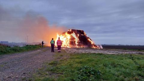 A large fire in the centre of the image surrounded by grass and flat open farm land. The fire is made up of rectangle shaped hay bales which have turned from a light yellow colour to a charred black. Flames can be seen coming out from the stack of hay and dark grey smoke is rising to sky on the left of the image. In front of the large fire is two people watching it. One in a yellow high visibility police jacket and another wearing an orange high visibility fire and rescue jacket. 