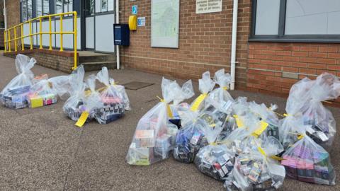 Approximately 20 clear bags filled with vapes and cigarettes on the floor in front of a police station. Each bag has a yellow ID tag.