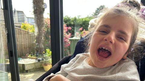 Edith sat in a wheelchair by a window looking at the camera with her hair tied back. She is wearing a grey top.