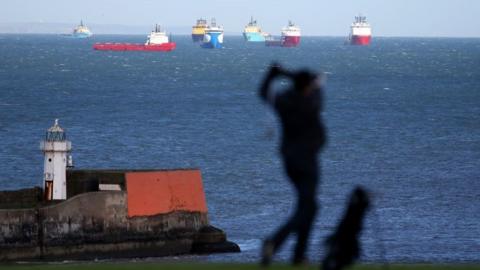 Silhouette of a golfer, with a lighthouse in the background, and offshore industry vessels in the sea.