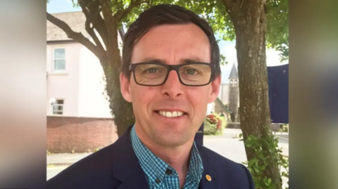 Darren Price yn edrych tuag at y camera gyda choed ac adeilad gwyn yn y cefndir. Mae ganddo lygaid glas, gwallt brown ac yn gwisgo sbectol.