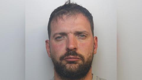 A mugshot of a man. He has a vacant expression, short brown hair and a beard and moustache