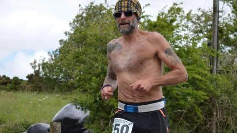 A topless runner with a checked hat and the number 50 on his trousers and trees and grass behind him.