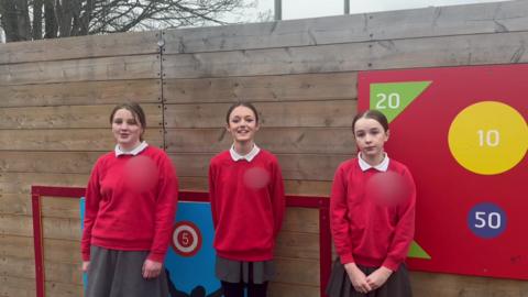 Three schoolgirls wearing red jumpers and grey skirts standing in front on a wooden panelled wall. On the wall, there are boards with numbers on them in colourful shapes.