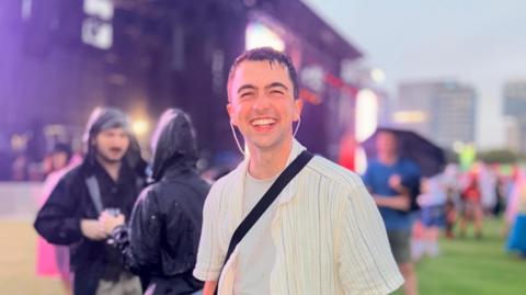 Alex Milsom is wearing a wet striped t-shirt and has wet hair. He is standing in a festival area in front of a stage with other people wearing rainproof jackets.
