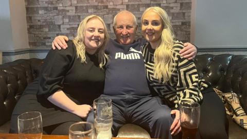 Faye (left) and Lisa Bramall sitting on a sofa in a bar, either side of their father Philip Bramall, who has his arms around them.