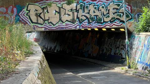 The image shows a dark underpass. There is graffiti on the walls outside. In the image the road curves to the left.