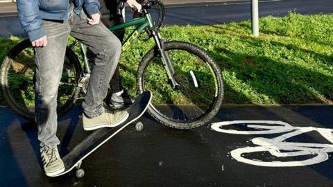A person on a skateboard from the waist down and another person with a bike. Both are on a cycle path.
