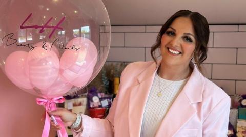 Lucy Howlett, a woman wearing a pink suit jacket is holding a clear balloon with her company logo on it containing small heart-shaped balloons inside it.