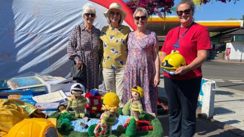 Four women stand behind a yarn display of firefighters rescuing someone from water