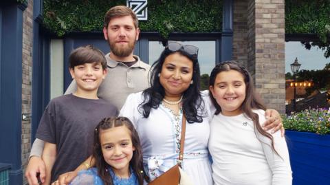 A family photo, a man with brown hair and beard and a woman with dark hair and sunglasses on her head smiling with their three children, all have brown hair.