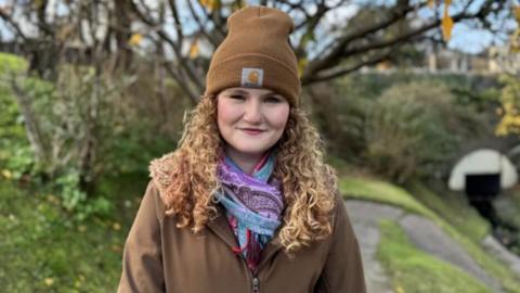 Millie is standing under trees with a stone bridge in the background. She has long curly strawberry blonde hair and is wearing a brown coat, brown hat and multi-coloured scarf.