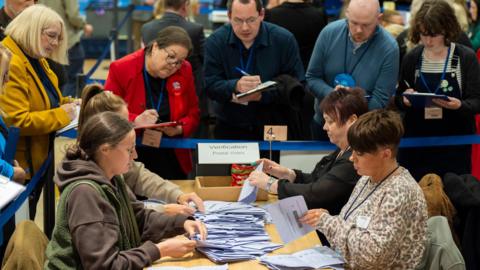 A pile of ballot papers are being counted on a table by four women. Behind them are representatives of the political parties, holding clipboards. 