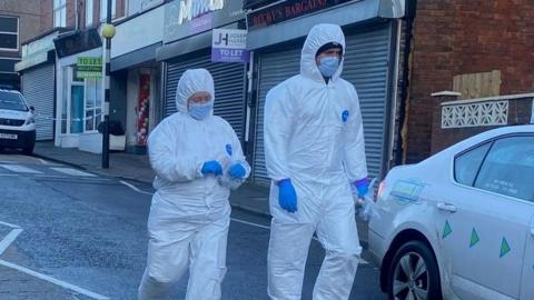 Two forensic officers dressed in white body suits and hoods with blue gloves on walk down a street.