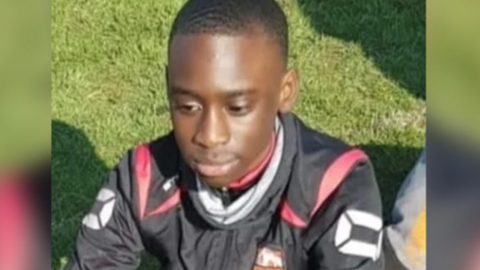 A mid-shot of a teenage boy in a black and red jacket sitting on some grass. He is looking down towards the ground. 