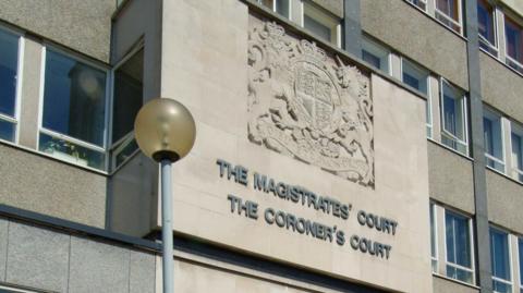 Exterior view of the stone facade of a building featuring the carved text "The Magistrates' Court" and "The Coroner's Court," with a royal coat of arms above and a streetlamp in the foreground.