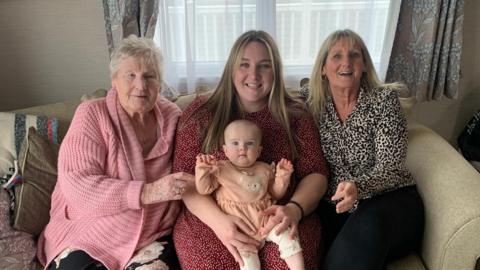 Four generations of women from one family sit on a sofa smiling at the camera