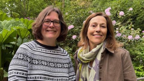 Two women stand in front of a hedge, smiling at the camera