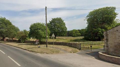 A grassy area behind a building. There are trees on the land. In the foreground is road.