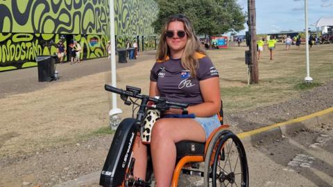 Martha Sharland pictured in a wheelchair wearing light blue denim short and a McLaren Formula One shirt that is grey and papaya. She has long brown hair and is wearing round sunglasses.