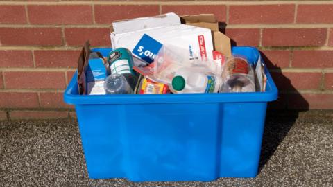 A blue recycling box on the ground outside house and against a brick wall. It is full of plastic bottles, tins and cardboard.