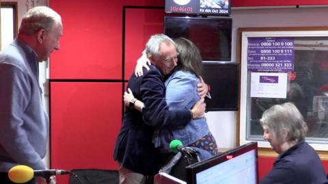 Dr Kevin Ives hugging Robyn Harris n in 鶹ҳ Radio Oxford's radio studio. Her parents are beside them. A digital clock on the wall shows the time.