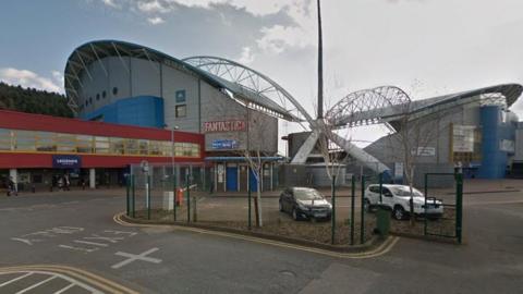 The outside of Huddersfield's John Smith's Stadium showing the back two of its arched stands, with an access road and car parking area in front.