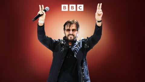Ringo Starr holds up his hands, holding a microphone.