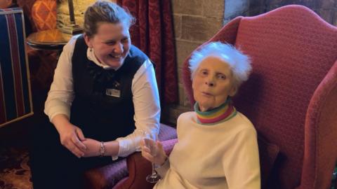 Molly Robinson and a female staff member are sitting on comfy red chairs in the castle. They are smiling. Molly, who has white hair and wears a white jumper with a rainbow-coloured collar, holds a glass of champagne.