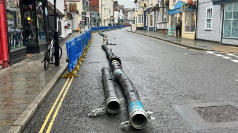 A large pipe seen on the road next to blue plastic fencing and with shops and buildings in the background