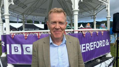 Adam Henson has swept back short blond hair. He is wearing a light blue shirt and a light brown tweed jacket. He is stood in front of a white band stand with BBC Hereford & Worcester brand draped across it.