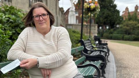 A woman wearing a white woollen jumper is sitting on a green park bench. She's holding a white sign, which is tied to the bench. There are buildings, a path and hanging baskets in the background.  
