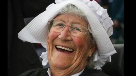 A smiling woman wearing glasses and a white headpiece that resembles a bonnet. Grey hair is visible beneath  the head covering and she has a dark top on. 
