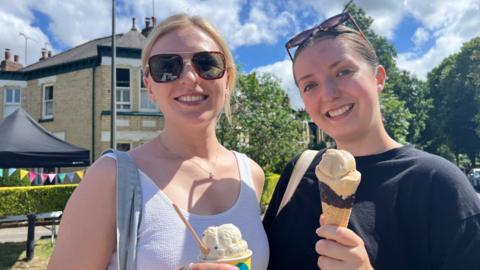 Two women smiling into the camera. The woman on the right has blond hair and sunglasses on. The woman on the right has brunette hair with sunglasses on her head. They are both holding ice cream. They are standing in front of a leafy avenue, with houses and a black marquee visible.