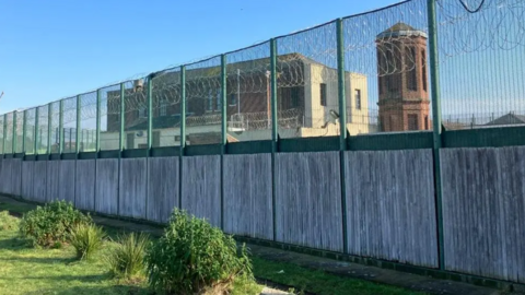 A tall wire fence on the perimeter of Haslar IRC, with a tall building and a brick tower inside the compound