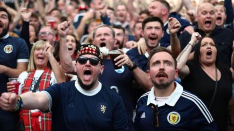Large crowd of football supporters wearing dark blue Scotland tops, gathered closely together in an indoor venue, many holding plastic cups of beer with arms raised, including a person in the foreground wearing a tartan hat.