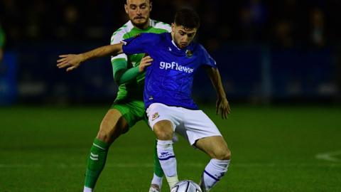 Aaron Henry shields the ball from a Yeovil Town player, wearing Wealdstone's blue kit with white shorts and socks