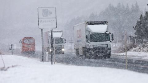 Lorries make their way along a snowy A9 with a sign for average speed cameras only just visible