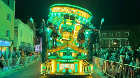 Big cart which is full of green and yellow LED lights shining. There's performers hanging off it. It's got the Renegades logo at the bottom. The cart is going through the street with people lining the street watching it.