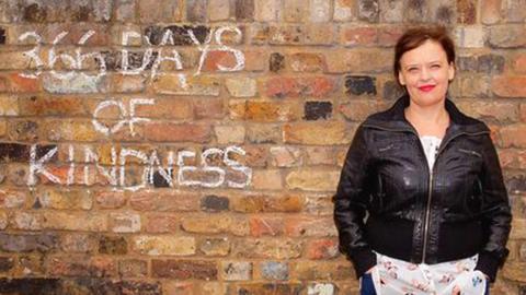 A woman stands against a wall with 366 days of kindness written on it