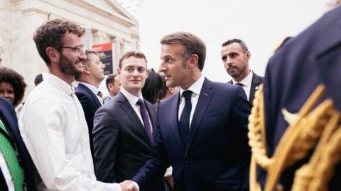 A man wearing a white long sleeved shirt shaking the hand of President Macron in a busy building. There are other men surrounding them in a line.