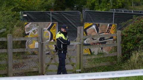 A man wears a black and hi-viz yellow jacket with black trousers. He wears a navy baseball cap. Behind him is a fence with a black sheet on it. protecting an area of green woodland.