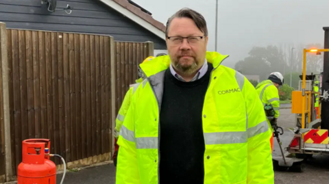 The image shows a bearded man in a hi-vis jacket looking at the camera. He is standing outside. There is a fence behind him to the left. To the right, there is a workman with a tool and machinery on the road.