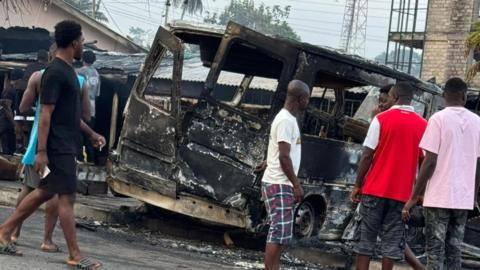 People stand in front of the remains of a burnt-out bus