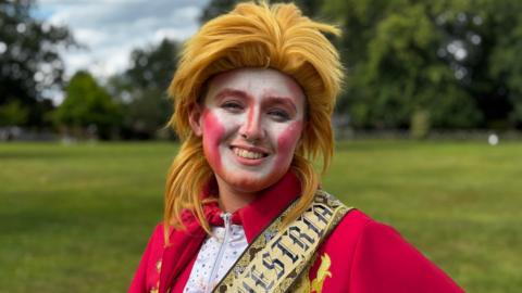 A drag performer in orange wig with their face painted white and red, wearing a gold sash with black writing that says Oswestry and a red and white zip-up Bowie style jumpsuit with rainbow sparkles.