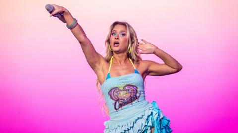 Zara Larsson performs on stage and she is holding a microphone outstretch to the crowd. She wears a blue sparkly dress.