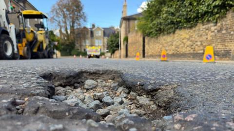 The photo is taken from the road level in a residential street. It shows a pothole in the foreground, around 8cm deep, where the asphalt surface has broken away, leaving course rubble below. In the background, machinery and highways vehicles have yellow lights flashing as they prepare to repair it.