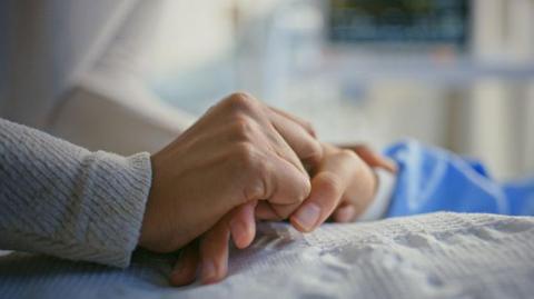 A person's hand holds the hand of a patient on a bed, with medical equipment in the background