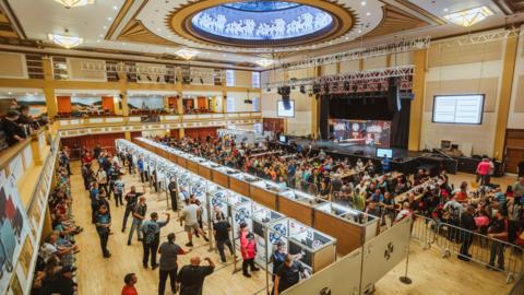 Interior of Bridlington Spa with rows of partitioned dart boards in the foreground in front of rows of desks with dozens of people sitting at them and a raised stage in the background.