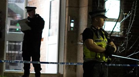 Police officers stand outside a flat. One is guarding a cordon, one is looking at a clipboard and two others are talking.
